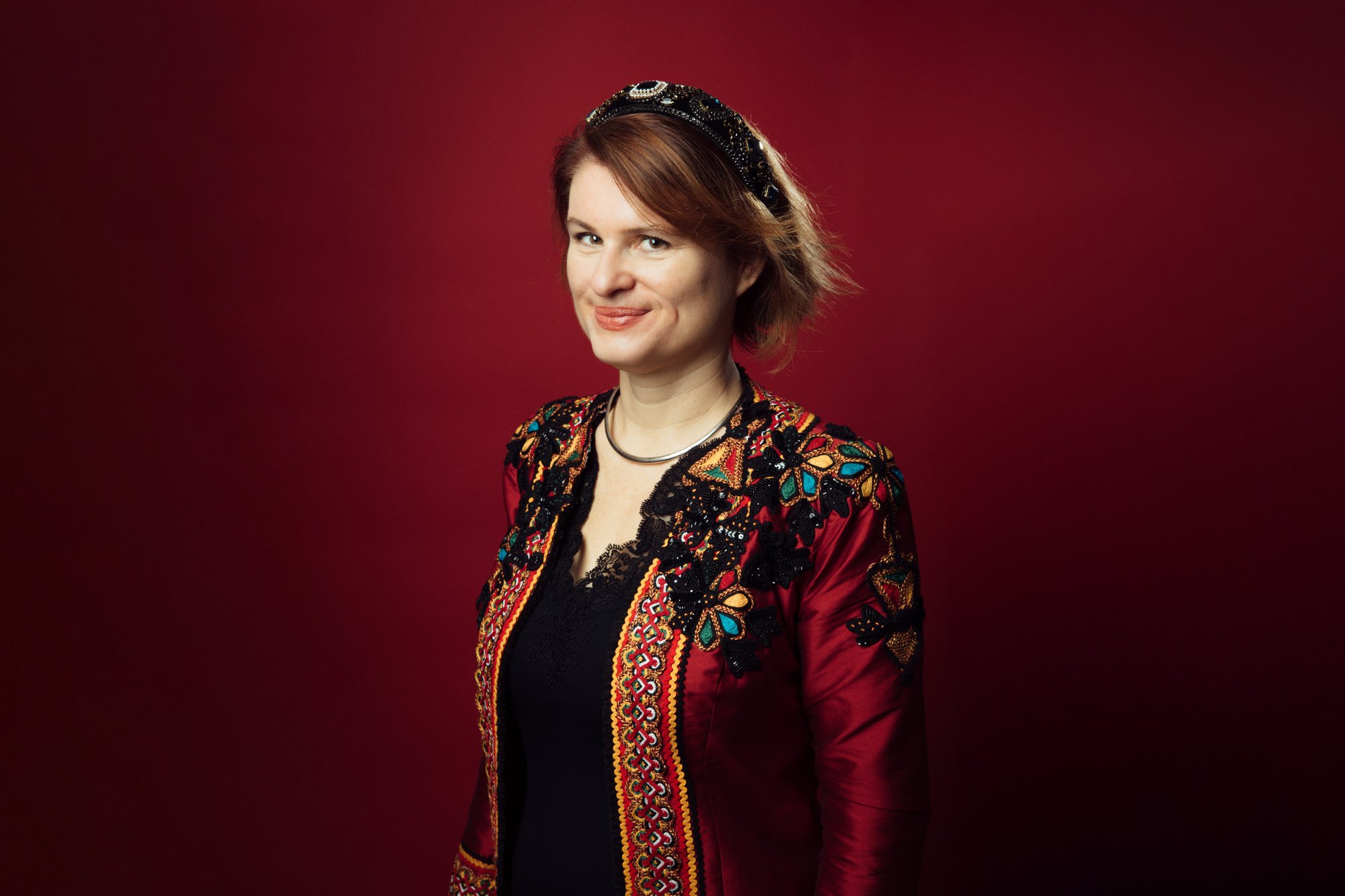 Portrait d’Audrey Alwett en veste rouge brodée et serre-tête, sur fond rouge